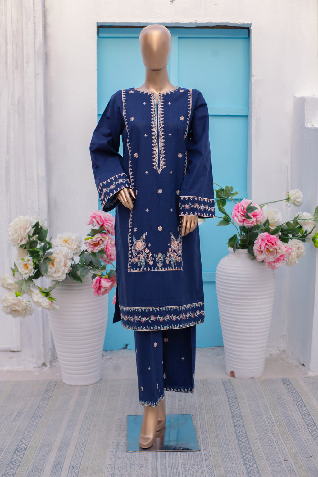 Mannequin wearing a blue embroidered outfit with floral arrangements in the background.