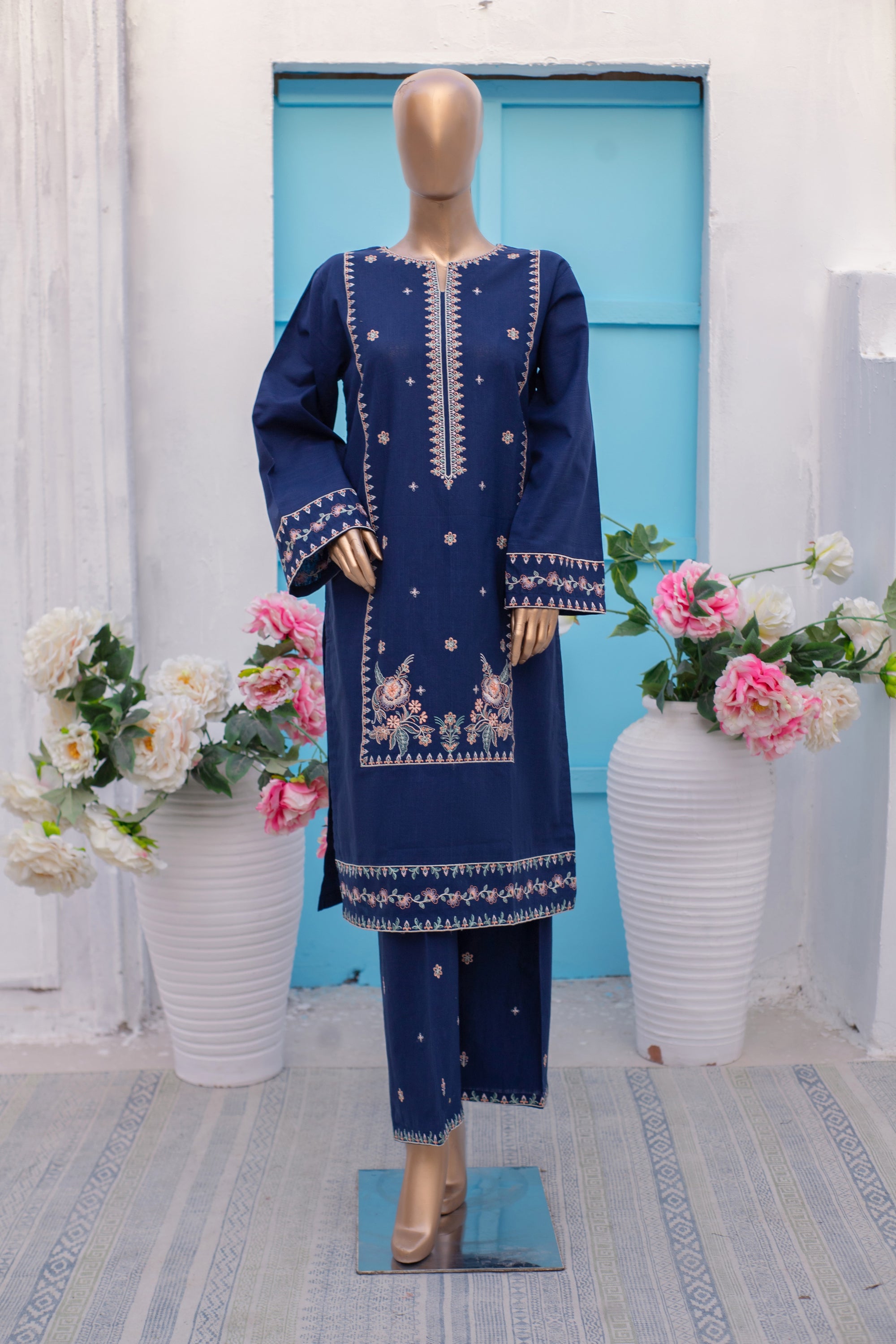 Mannequin wearing a blue embroidered outfit with floral arrangements in the background.